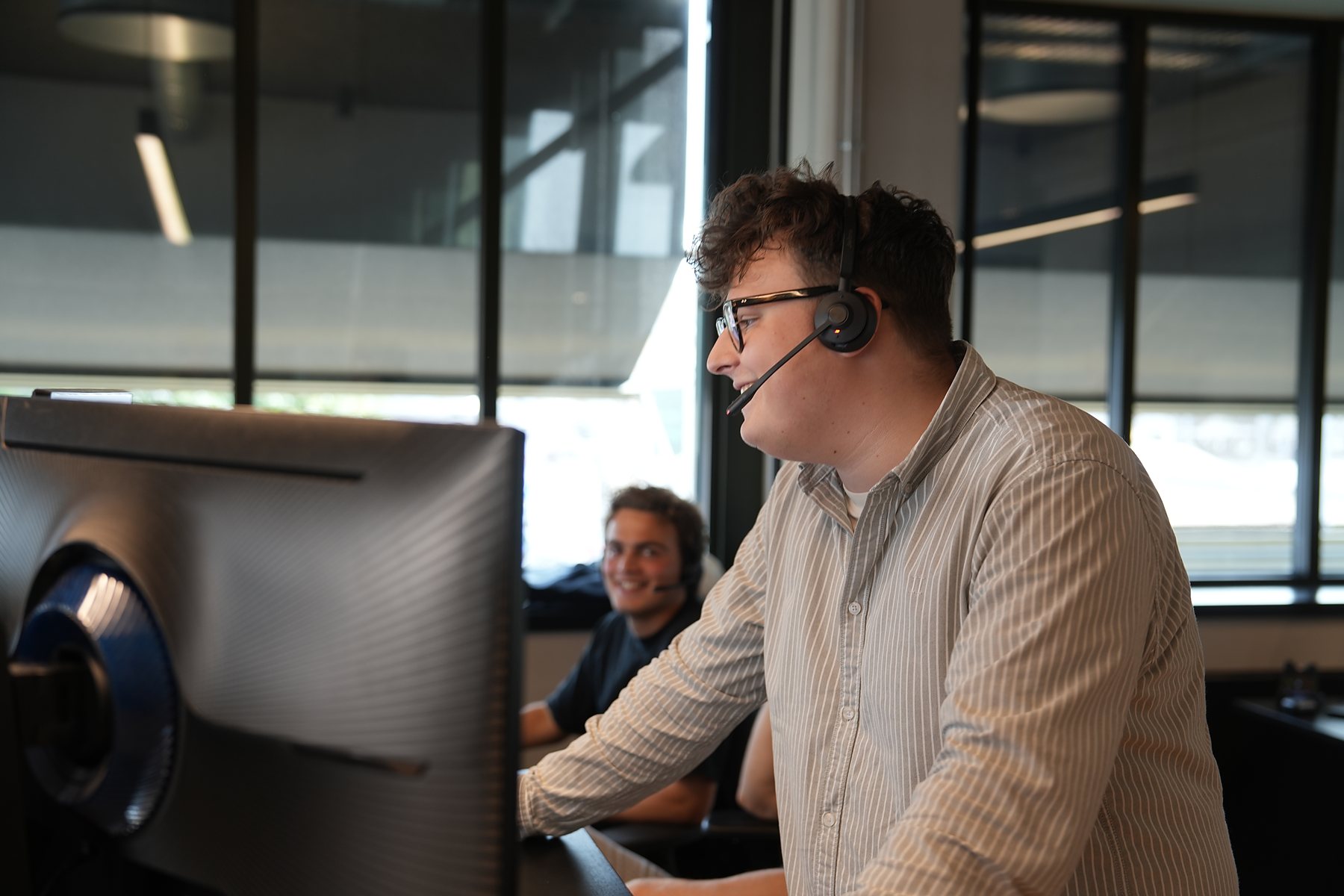 SDR med briller og headset smiler midt i et opkald, kollega griner bagved